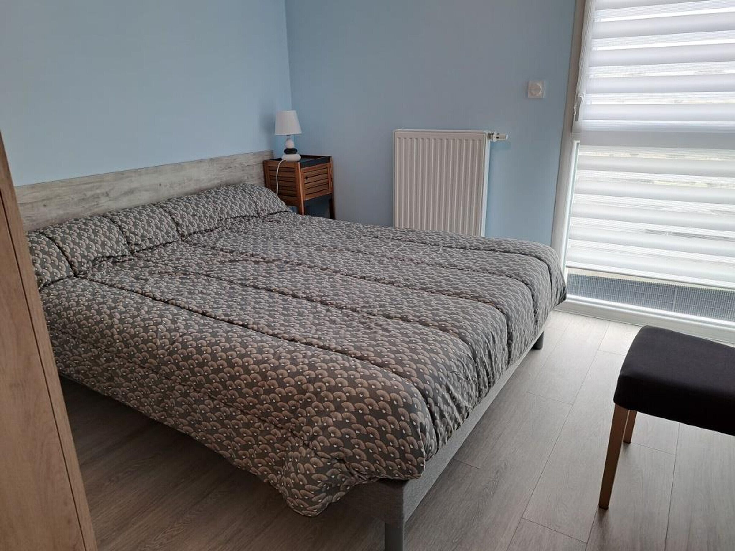 Une chambre à coucher comprenant un lit, une table de chevet, une lampe, un radiateur et une fenêtre avec des stores.