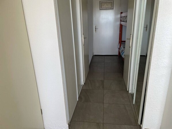 A narrow hallway with white walls and a tiled floor, leading to a door with a small window.