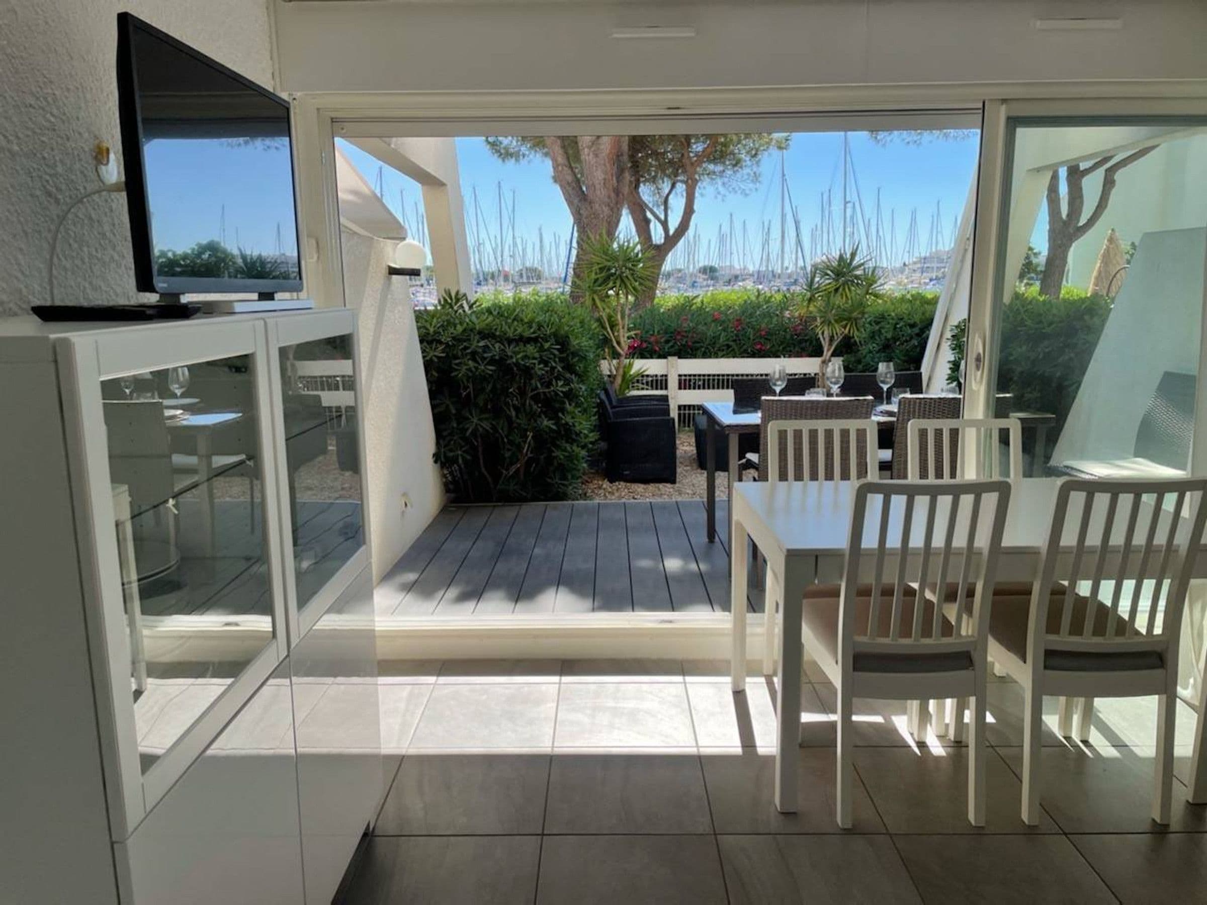 A patio area with a dining table and chairs, a television mounted on the wall, and a view of the sea with sailboats.