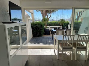 A patio area with a dining table and chairs, a television mounted on the wall, and a view of the sea with sailboats. - Ground Floor Apartment with Terrace, Garden, Pool & Tennis, 50m from Beach in Port Camargue (Port Camargue)