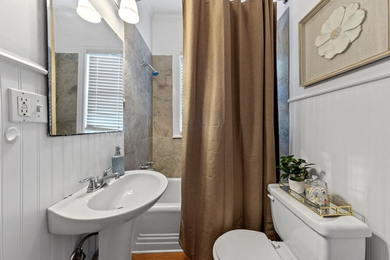 Une salle de bain avec un lavabo blanc, une baignoire et des toilettes. On y trouve un rideau de douche, un cadre décoratif avec des fleurs et une plante en pot posée sur les toilettes.