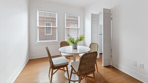 Una habitación con una mesa redonda, cuatro sillas de mimbre, una planta en maceta y dos ventanas con persianas.