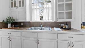 Una cocina con armarios blancos, un lavamanos doble y una ventana con cortinas translúcidas.