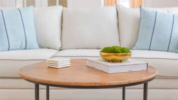 A living room with a white sofa, a round wooden coffee table, a bowl with a green plant, and a stack of books.