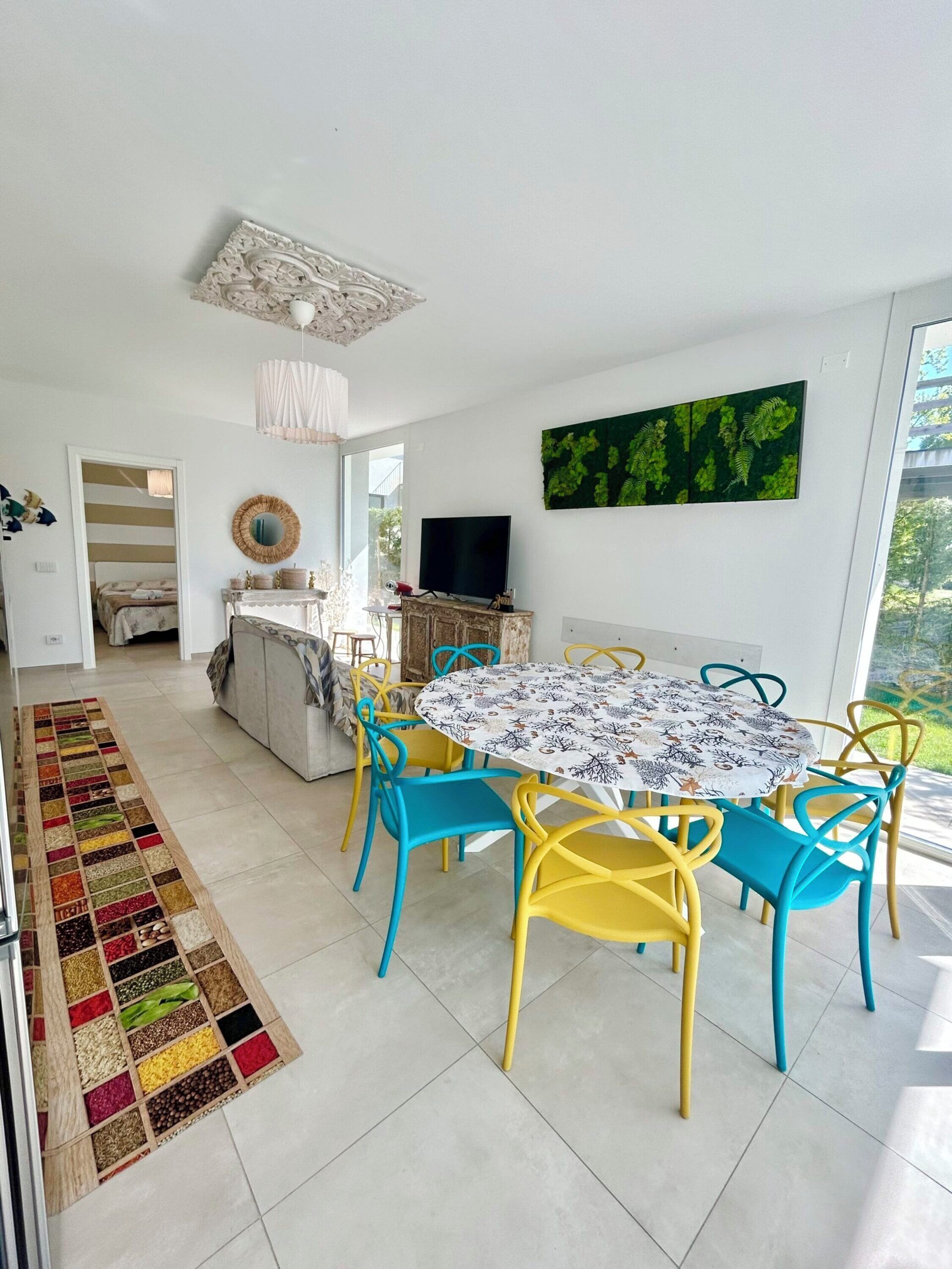 A modern living room with a round table and colorful chairs, a television, and a large window with a view of greenery.