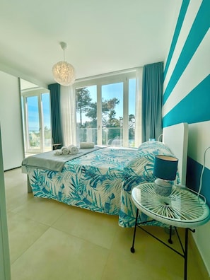 A modern bedroom with a large bed, a bedside table, a lamp, and a view of the outdoors through large windows. - Golden Hour Retreat Lagoon View Villa With Pool (Rosolina)