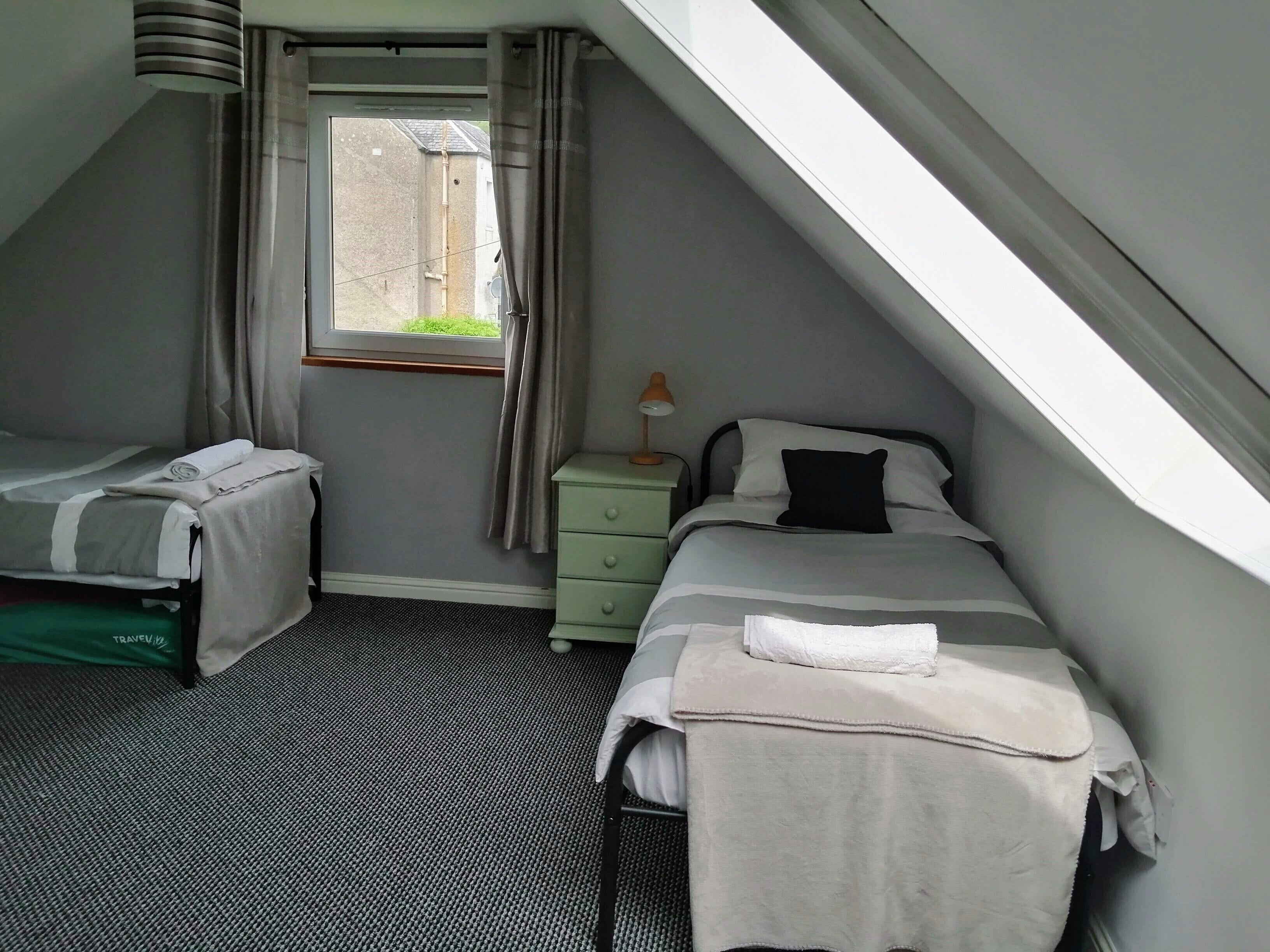A small, tidy room with a sloped ceiling, a single bed with a white blanket, a bedside table with a lamp, and a window with curtains.