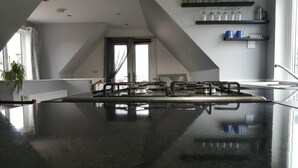 Une cuisine avec un plan de travail noir, une cuisinière à gaz et une fenêtre avec des rideaux. On y trouve des étagères avec de la verrerie et une plante sur le côté gauche.