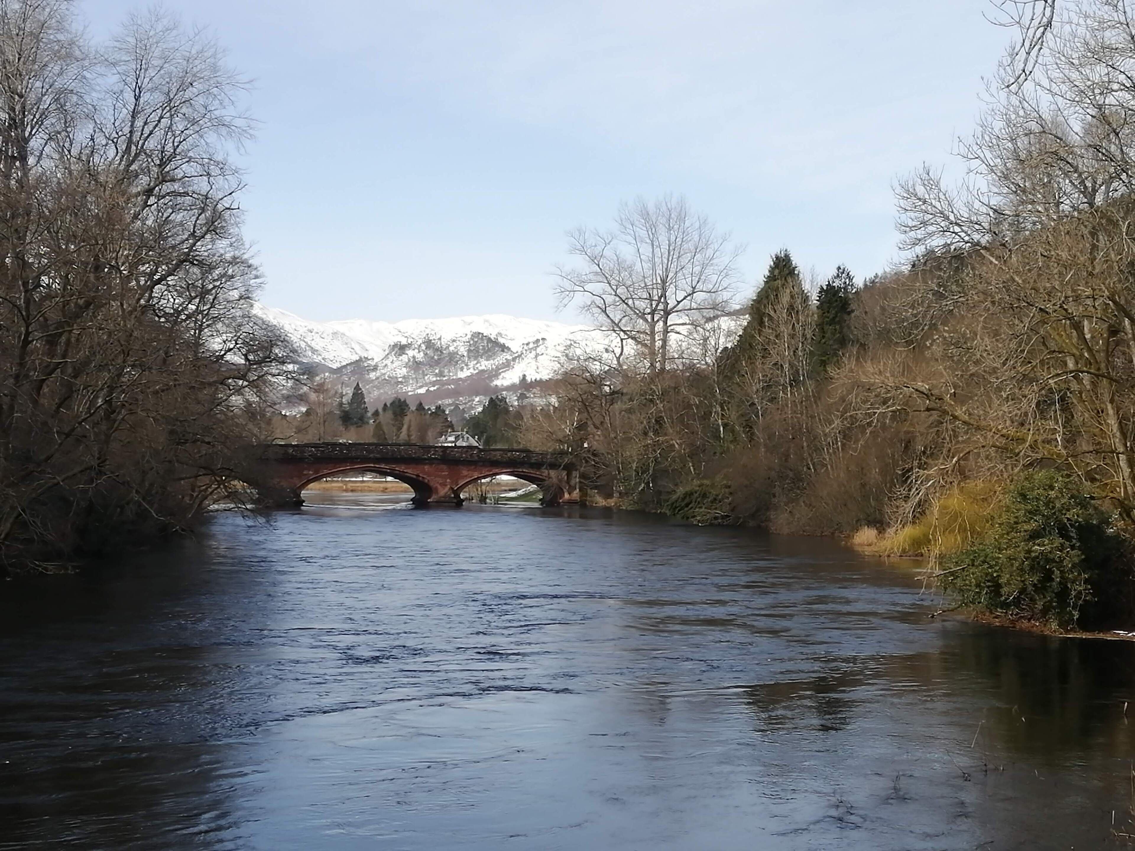 Une rivière traversée par un pont de pierre, bordée d’arbres sans feuilles et avec une chaîne de montagnes en arrière-plan.