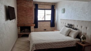 Een slaapkamer met een bed, een metselwerk schoorsteen, een raam met gordijnen en een aan de muur bevestigde televisie.