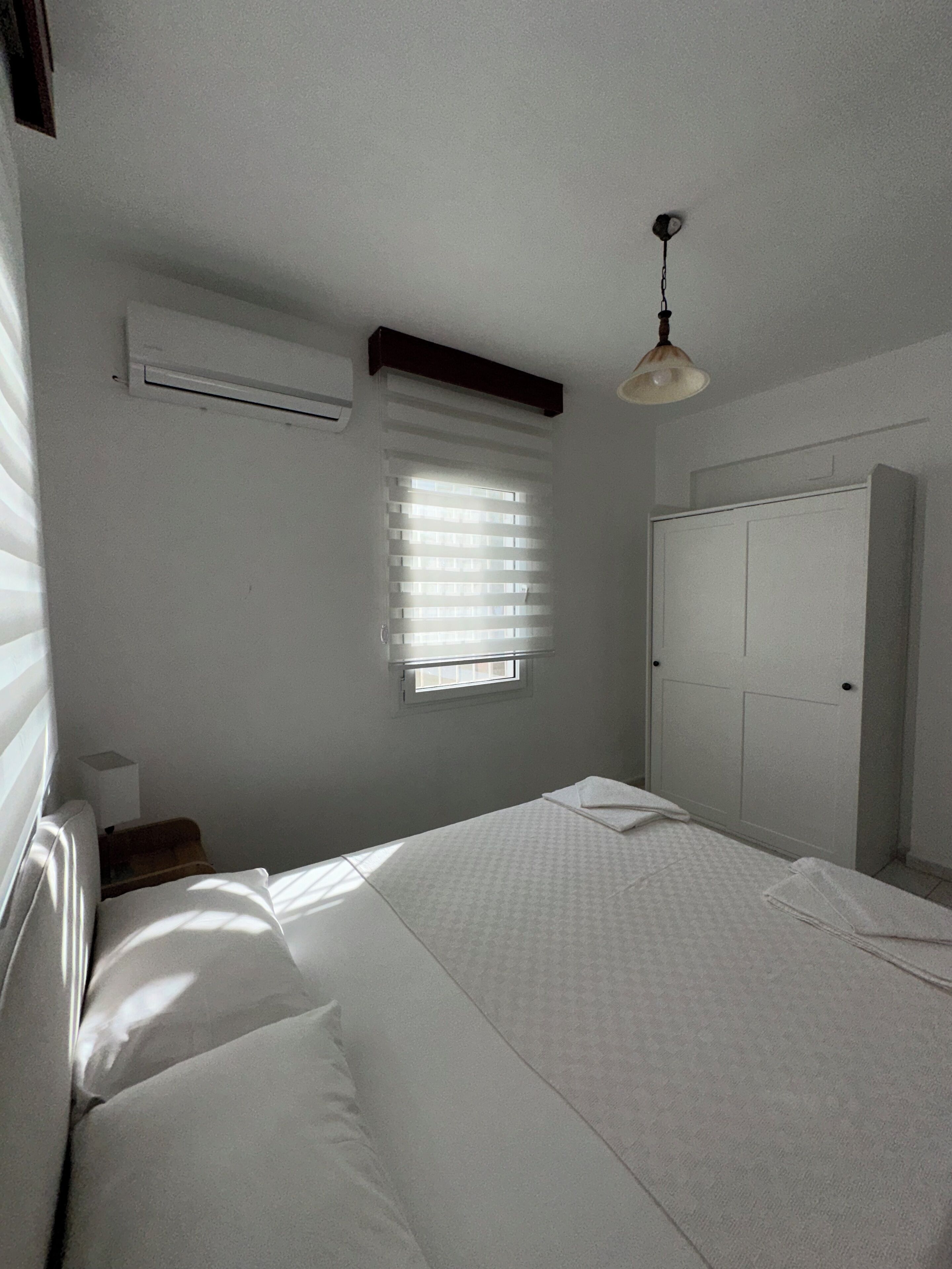 Un dormitorio con cama, ventana con persianas, equipo de aire acondicionado, armario y lámpara colgante.