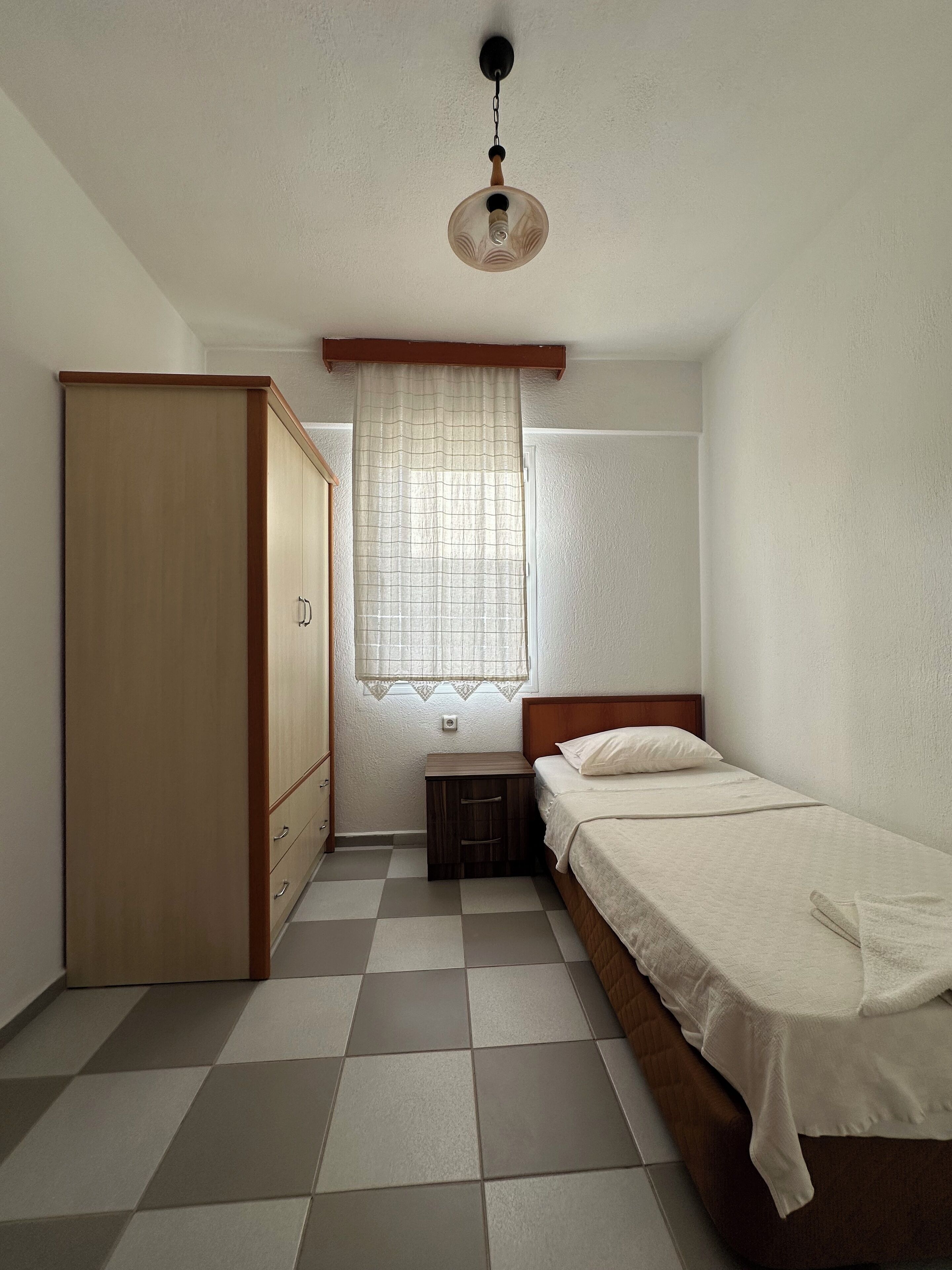 Um quarto com uma cama, guarda-roupa, mesa de cabeceira, janela com cortinas e luminária no teto.