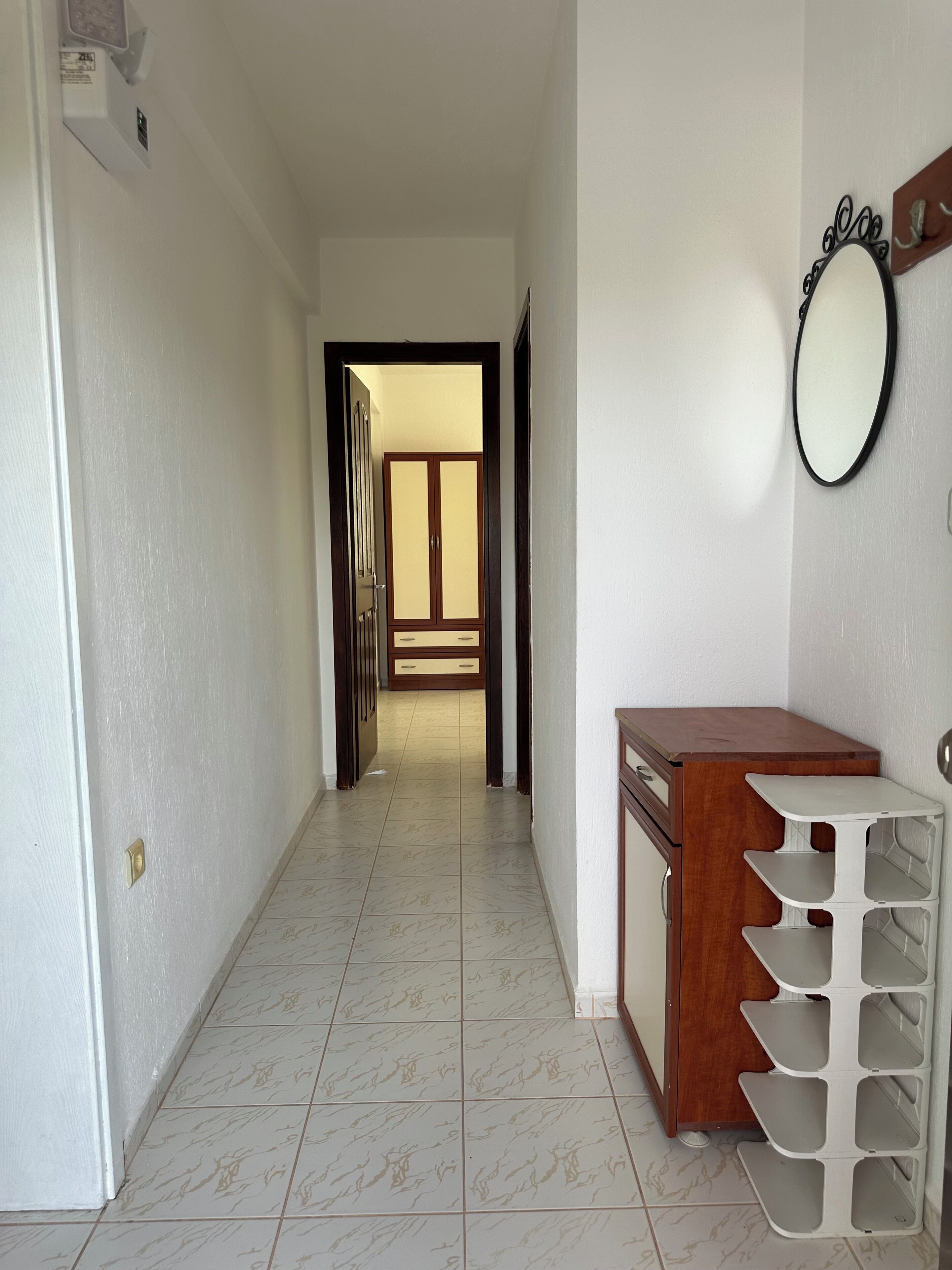 A hallway with a wooden cabinet and a white shelf unit, leading to a room with a wardrobe and a mirror on the wall.