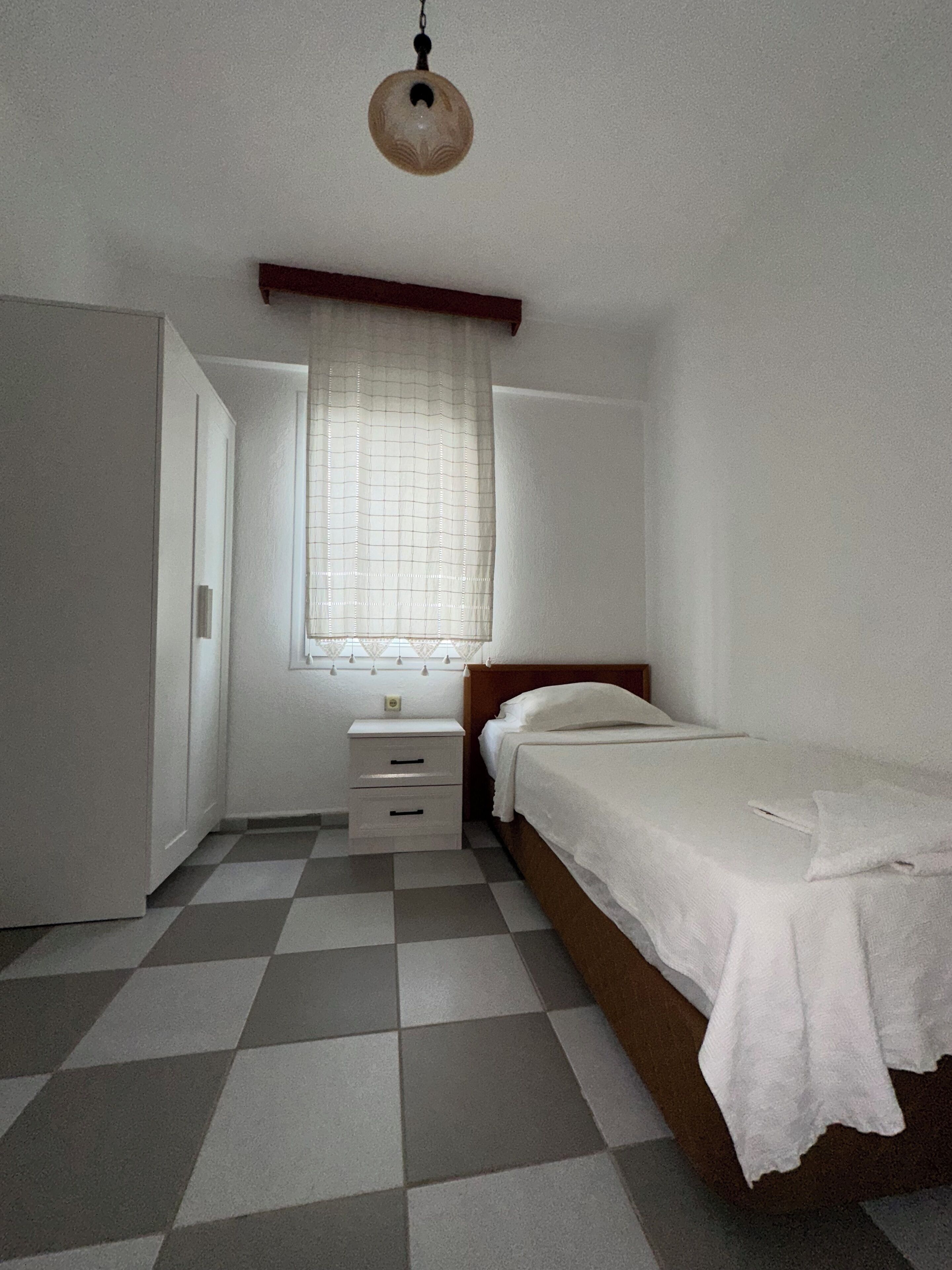 A small, clean room with a checkered floor, a single bed, a nightstand, and a window with a curtain.