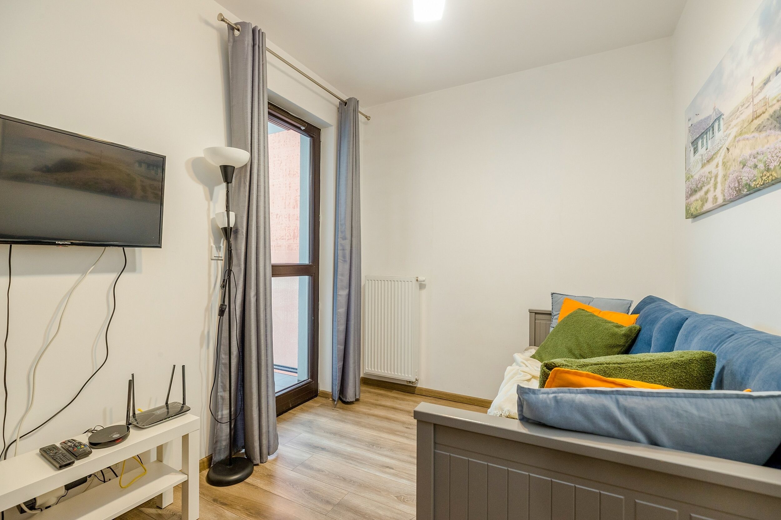 Una sala de estar moderna con un televisor de pantalla plana, una consola blanca con dispositivos electrónicos, un sofá azul con almohadones coloridos y un gran ventanal con cortinas.