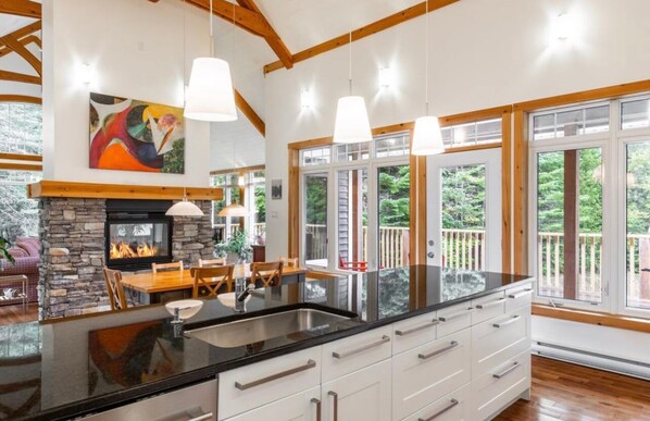 A modern kitchen with a large island, stone fireplace, and large windows offering views of the outdoors. - Spa and Lakeside | La Luz del Dia (Saint-Didace)