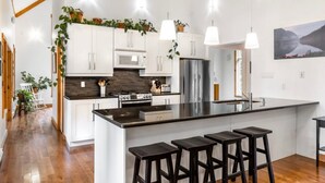 A modern kitchen with white cabinets, a central island with bar stools, stainless steel appliances, and hanging plants.