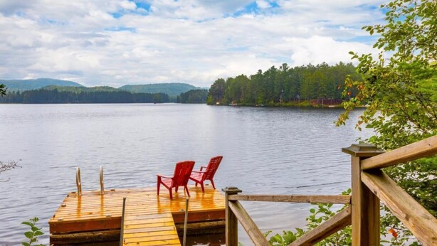 A wooden dock with red chairs overlooking a calm lake, surrounded by trees and distant hills.