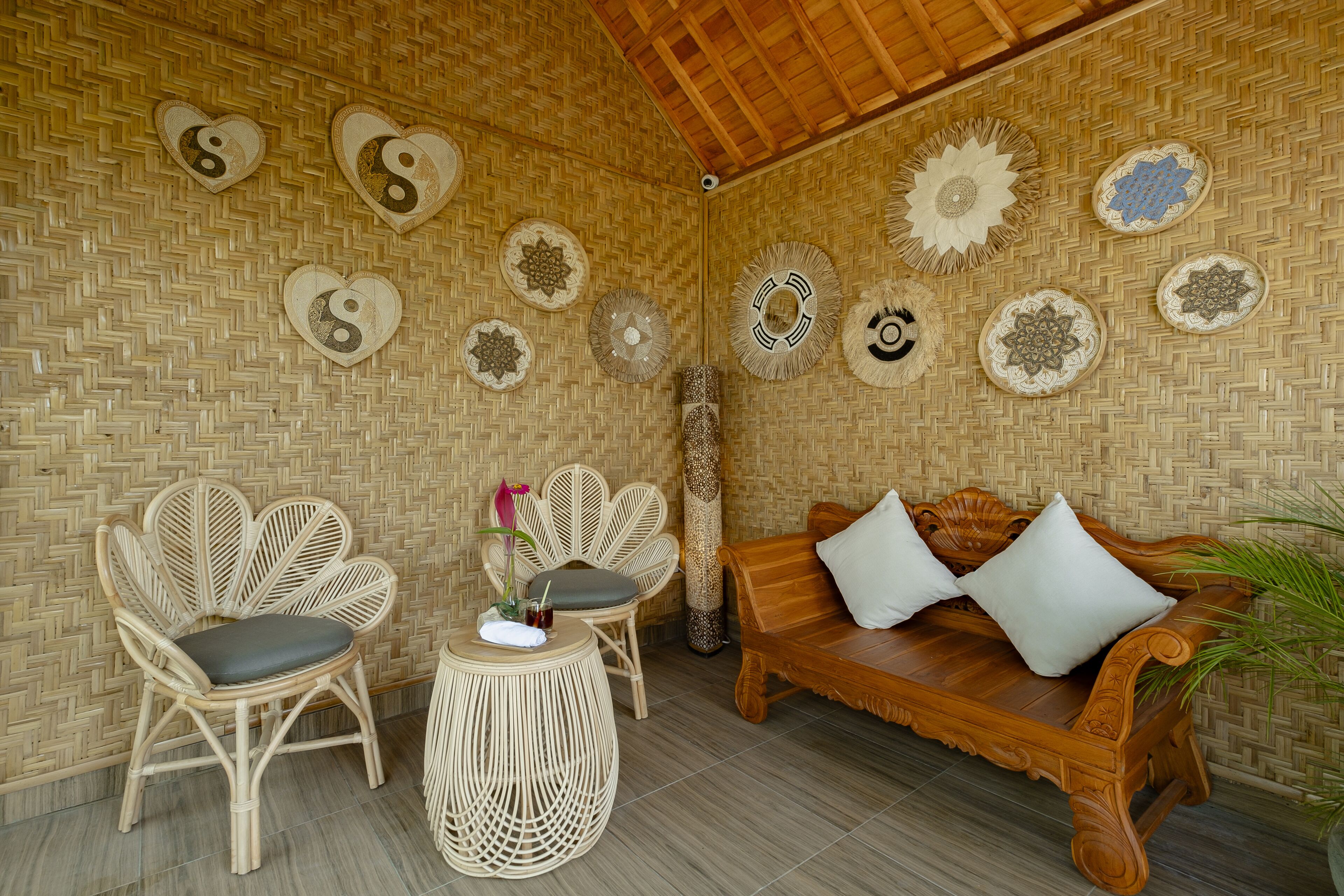 A room with a woven bamboo wall, a wooden sofa with cushions, two wicker chairs, and a small round table with a flower and a drink.