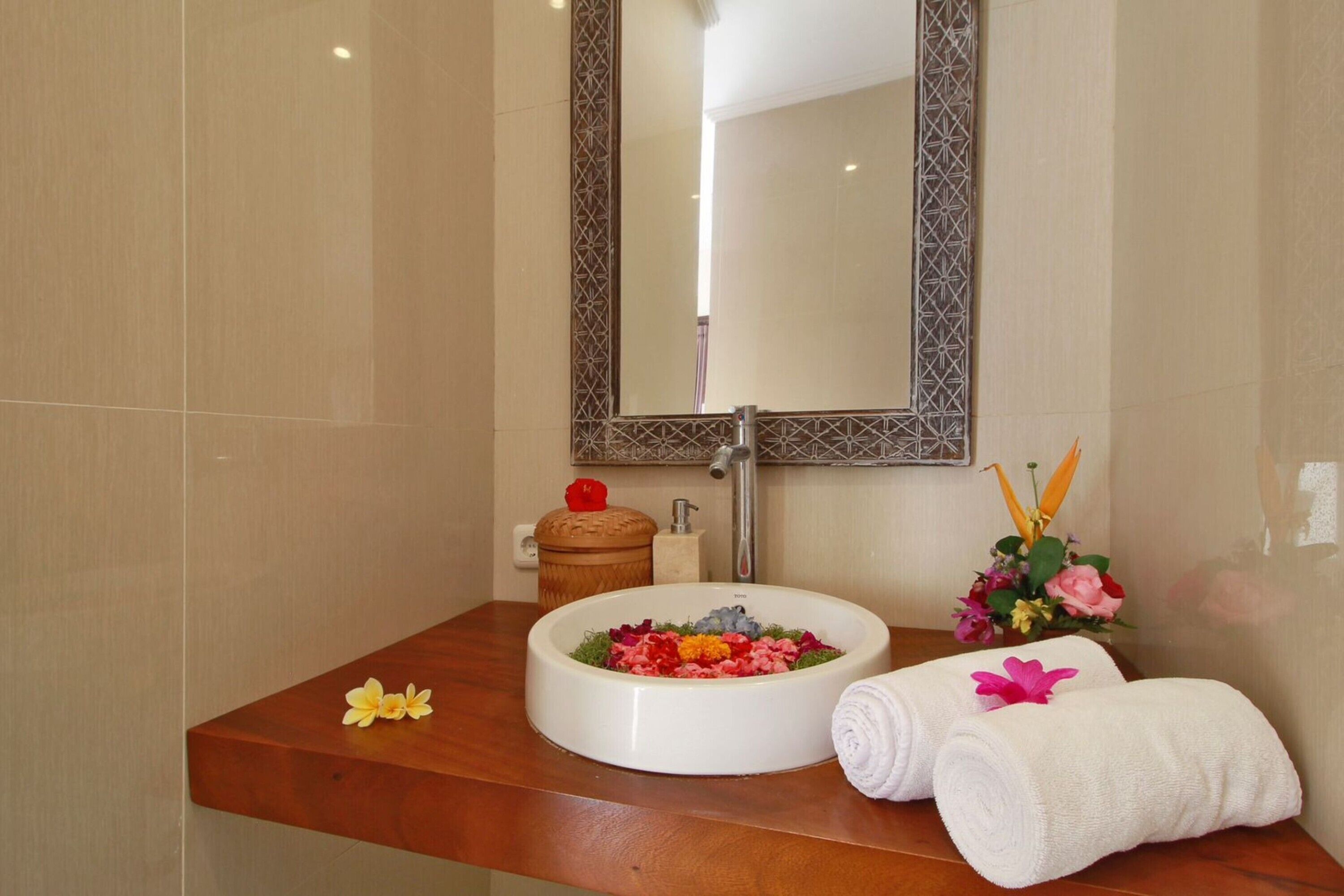 A bathroom with a wooden vanity, a white sink with a decorative bowl, a mirror with a geometric frame, and towels and flowers on the counter.