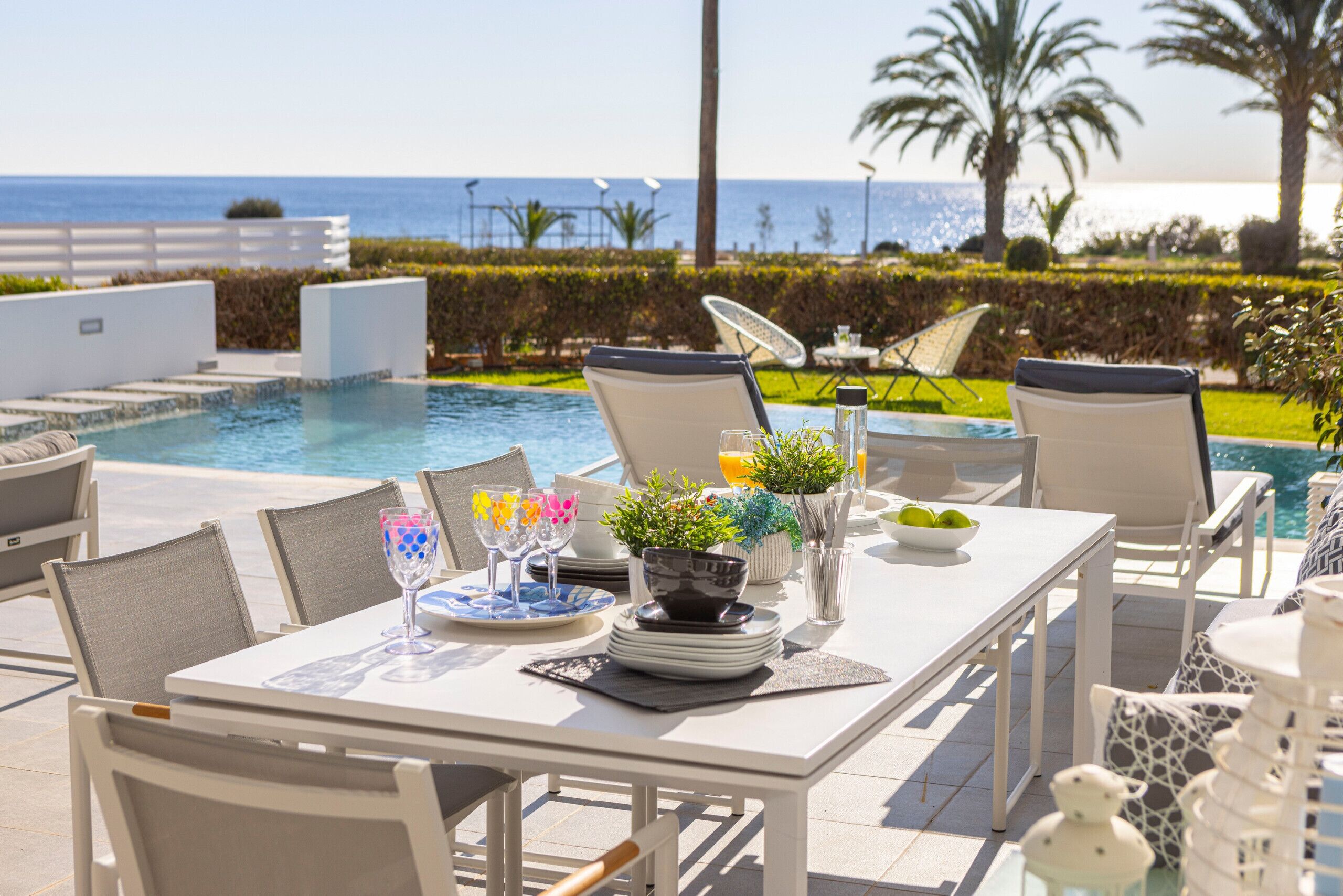 Un área de comedor al aire libre con vista a la piscina, muebles blancos y una mesa servida con platos, vasos y un cuenco de fruta.