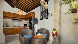 Een keuken met houten kastjes, een koelkast en een magnetron. Er staan rieten stoelen met kussens en een vaas met bloemen.