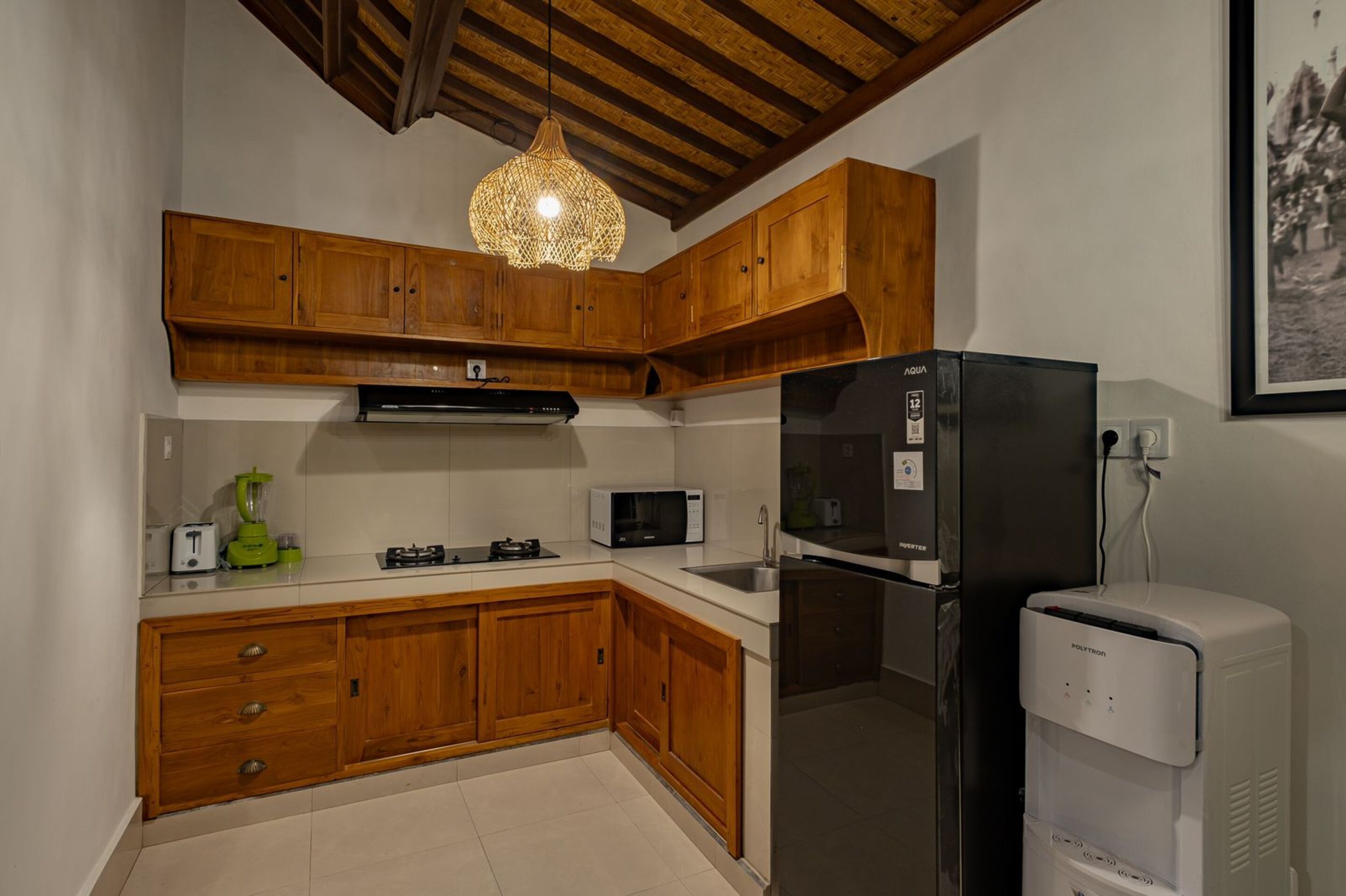 Una cocina con armarios de madera, un refrigerador negro, un microondas, un dispensador de agua y una lámpara colgante.