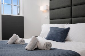 A neatly made bed with rolled towels, a grey headboard, and a window with blinds.