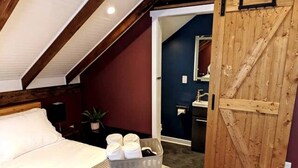 A bedroom with a bed, a bedside table with a lamp, a basket of toiletries, and a wooden door leading to a bathroom. - Walkable Elmwood Cottage, No Chores 2Bath 2BR 2Car (Buffalo)