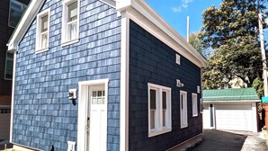 A two-story house with a dark blue exterior and white trim. It features a front door, two windows on the first floor, and a garage with a green roof. - Walkable Elmwood Cottage, No Chores 2Bath 2BR 2Car (Buffalo)