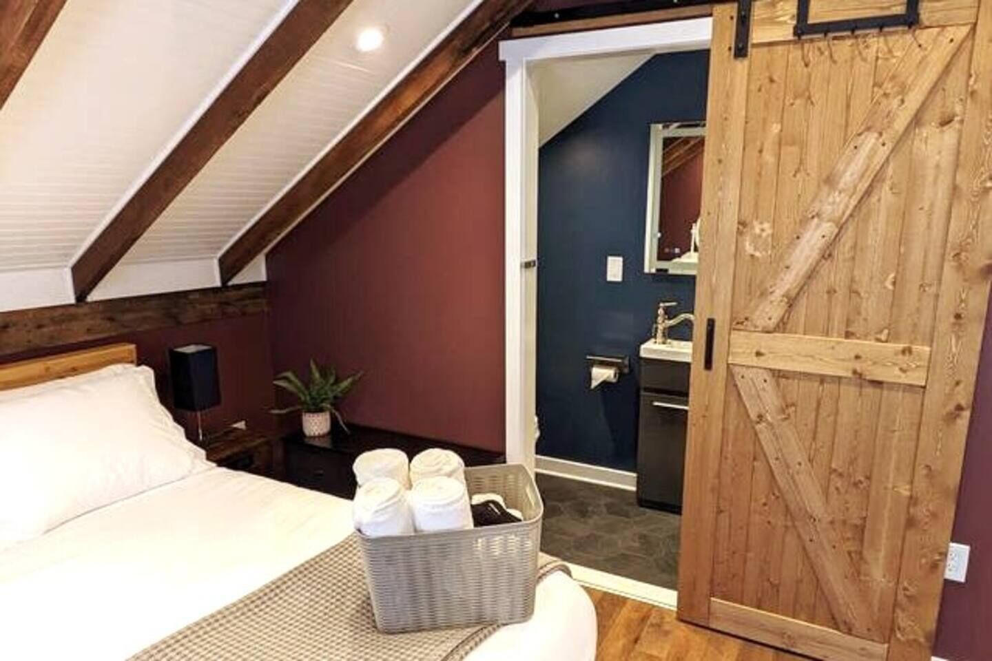 A bedroom with a bed, a bedside table with a lamp, a plant, and a basket of toiletries.