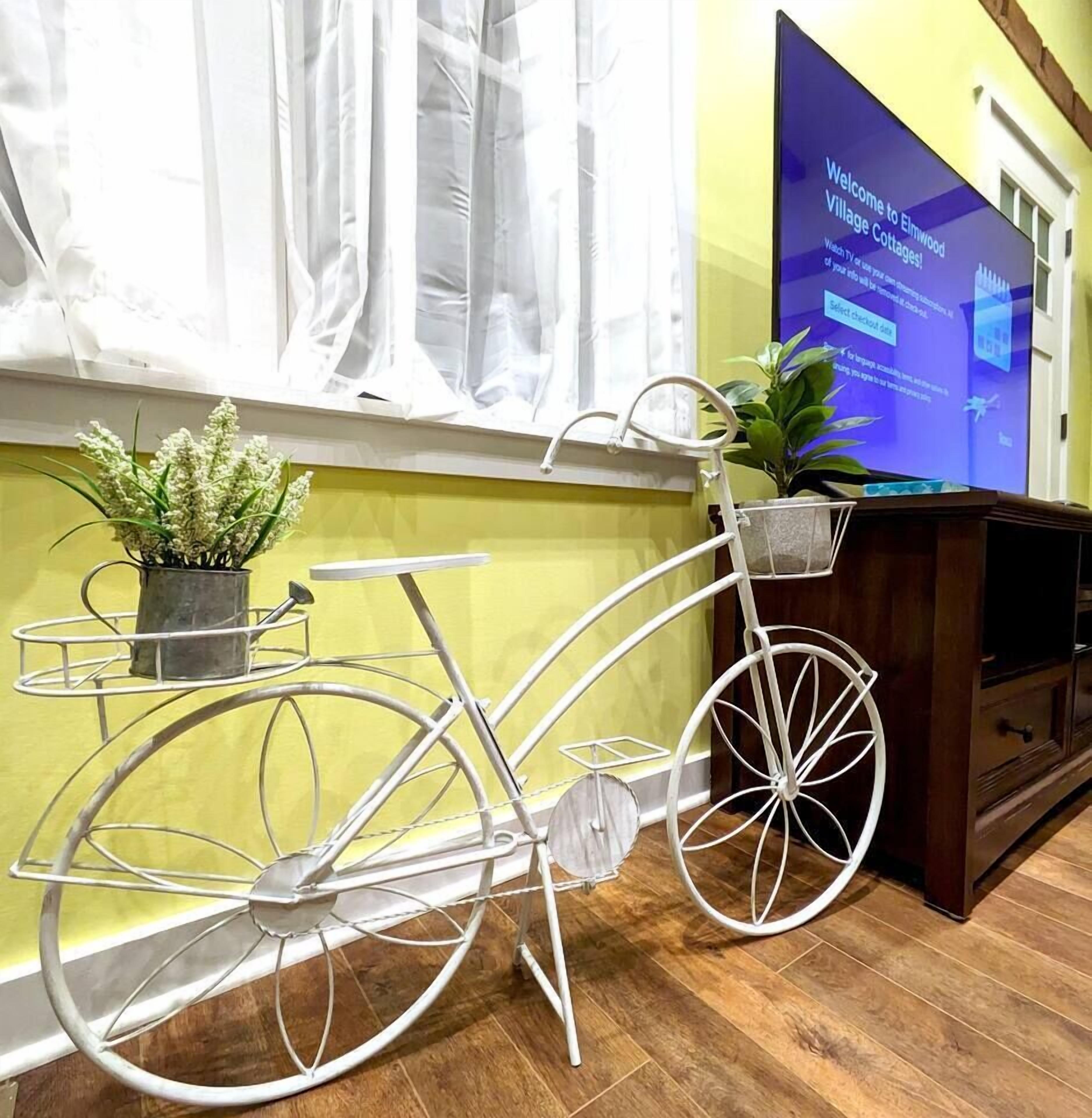 Una bicicleta blanca con una cesta y una pequeña planta a su lado, una pantalla de televisión mostrando un mensaje de bienvenida, y un suelo de madera.