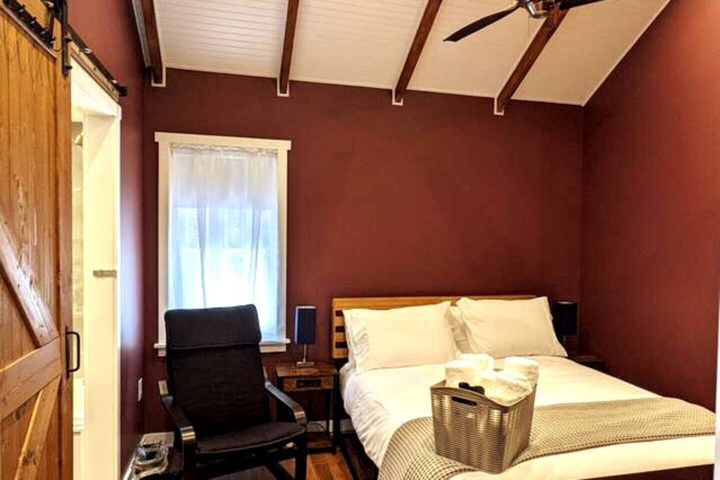 A bedroom with a bed, a chair, a nightstand, a window with curtains, a ceiling fan, and a wooden door.