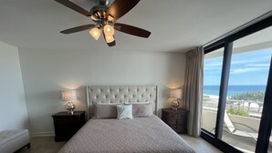 A bedroom with a large bed, two bedside tables with lamps, a ceiling fan, and a view of the ocean.