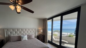 A bedroom with a large bed, a ceiling fan, and a view of the ocean through a sliding glass door.