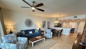 A spacious living room with a sofa, two patterned armchairs, a coffee table, and a ceiling fan. A kitchen with a dining area is visible in the background.