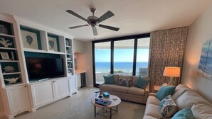 A living room with a television, sofa, and coffee table. There is a ceiling fan and a view of the ocean through large windows. - Gulf Front Condo With Stunning Views! (Pensacola)