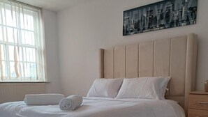 Ein ordentlich bezogenes Bett mit weißen Leinen und einem zusammengerollten Handtuch, ein Nachttisch aus Holz mit einer Lampe und ein Stadtbild an der Wand.