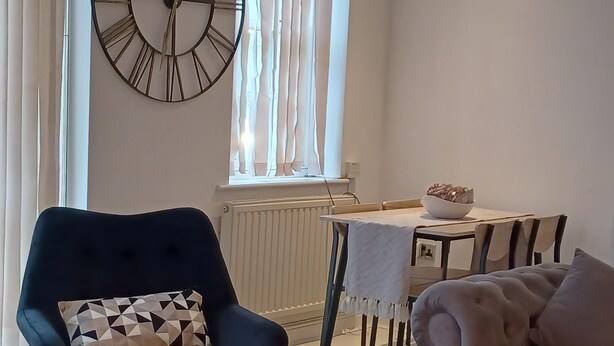 Ein modernes Wohnzimmer mit einem dunkelblauen Sessel, einem beigen Sofa, einem Esstisch mit weißem Tischtuch und einer großen Wanduhr.