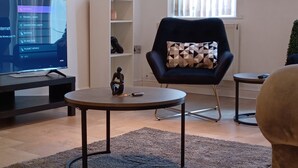 Ein modernes Wohnzimmer mit einem Flachbildfernseher, einem schwarzen Sessel mit geometrischem Kissen, einem kleinen Beistelltisch und einem Bücherregal mit Dekorationselementen.