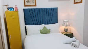 A bed with a blue headboard, white pillows, and a green accent pillow. There is a yellow wardrobe to the left and a bedside table with a lamp and a folded towel. - Surebillionaire Home Deluxe Queen Studio (London)