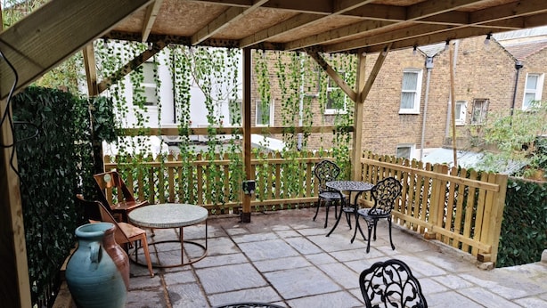 A covered patio area with stone flooring, wooden furniture, and a view of a brick building. - Surebillionaire Home Deluxe Queen Studio (London)