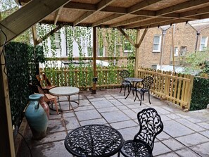 A covered patio area with stone flooring, wooden furniture, and a view of a brick building. - Surebillionaire Home Deluxe Queen Studio (London)