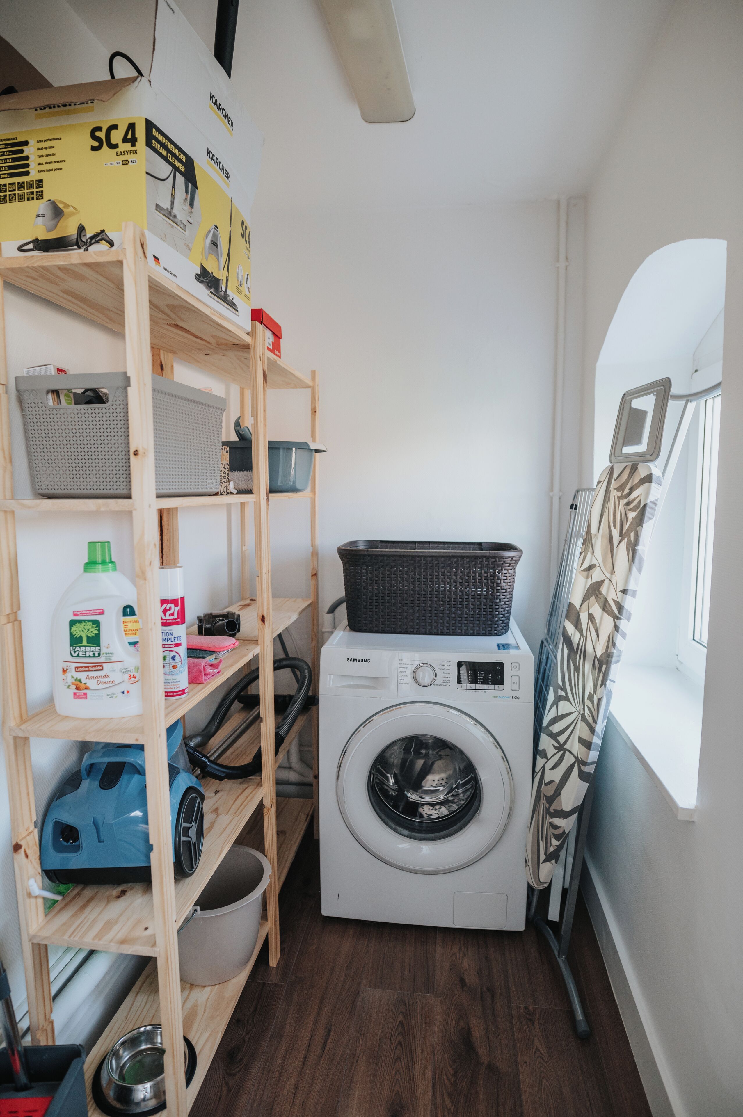 A laundry room with a washing machine, shelves with cleaning supplies, a basket, and a window with a curtain.