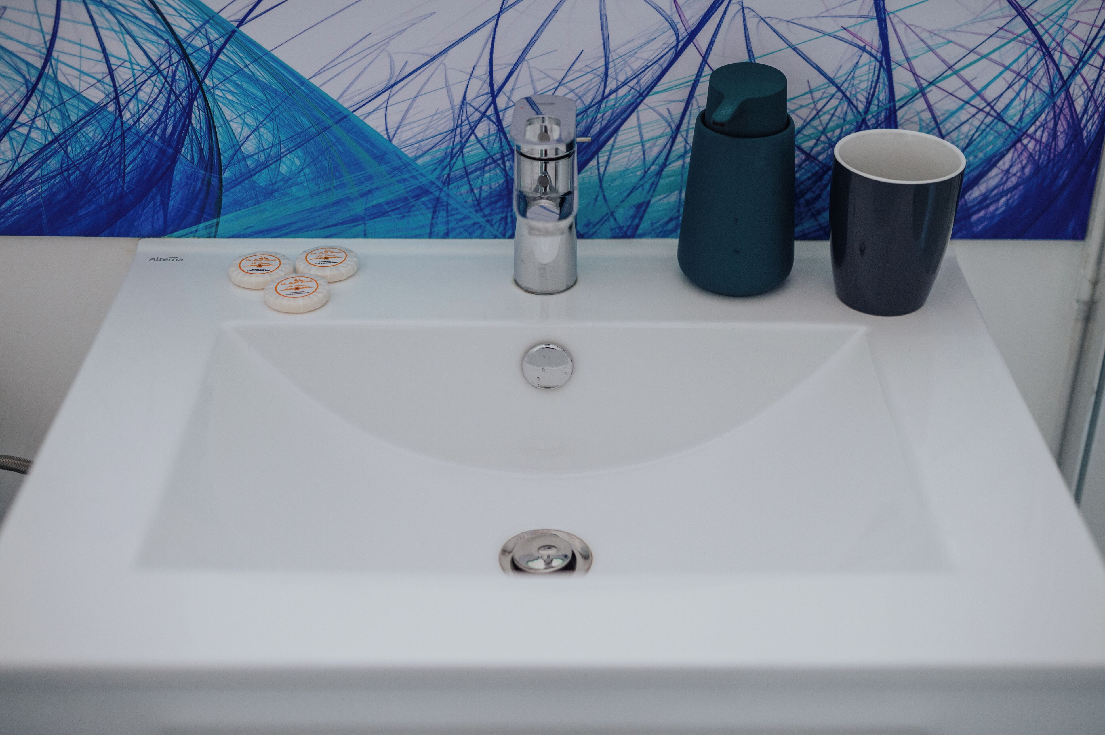 Un lavamanos blanco con un grifo moderno, una botella de agua azul turquesa y una taza oscura sobre una encimera blanca.