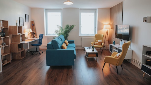 A modern living room with a blue sofa, yellow armchairs, a wooden floor, and a large window providing natural light. - Le Rêve Bleu (Thionville)