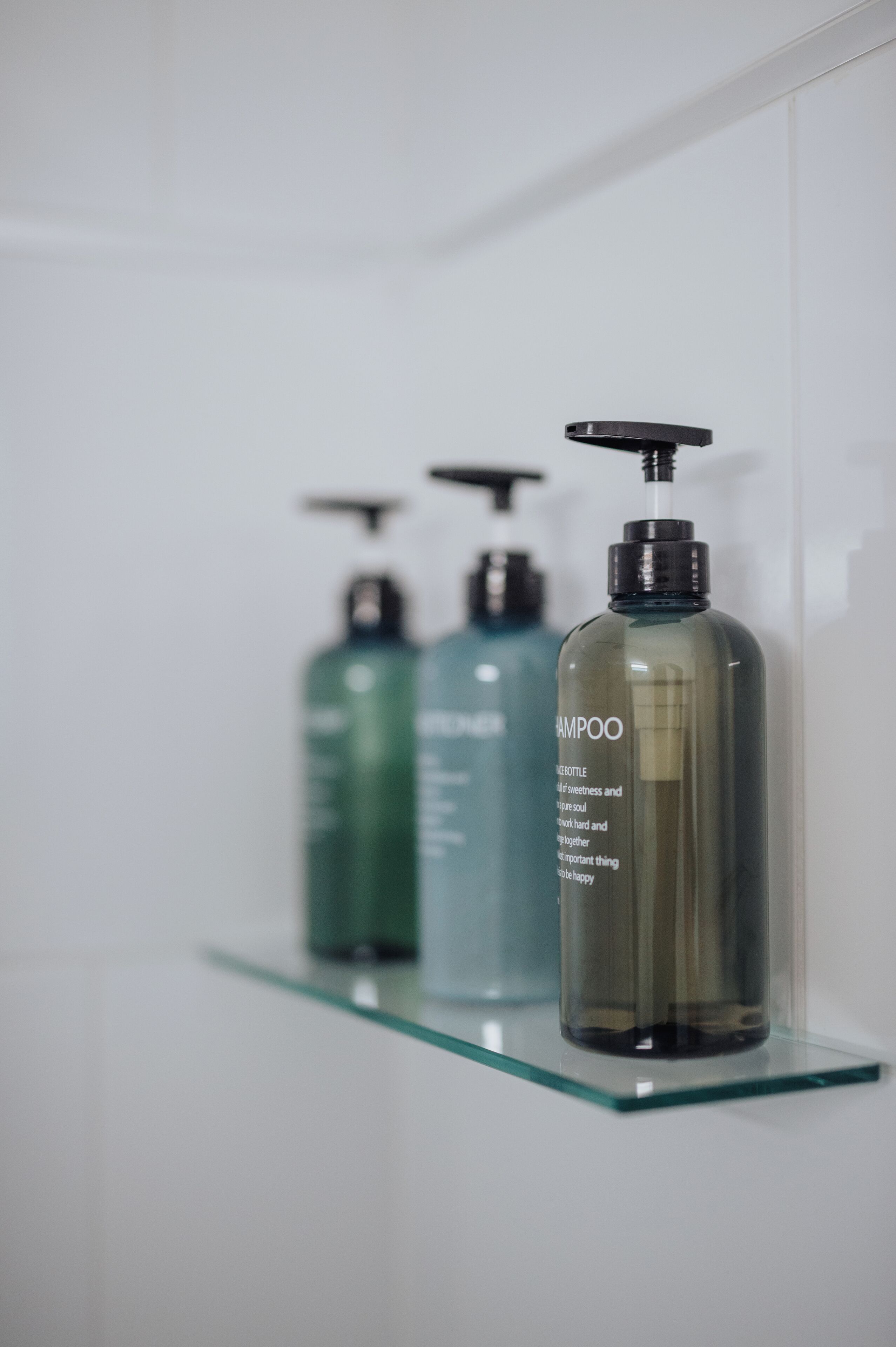 Three bottles of shampoo on a glass shelf against a white wall.