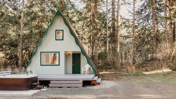 A small, triangular wooden cabin with a porch and a hot tub, surrounded by trees. - 54sl - Hot Tub - Pets Ok - Sleeps 4 2 Bedroom Cabin by RedAwning (Deming)