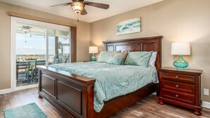 A bedroom with a large bed, wooden headboard, two bedside tables with lamps, and a view through a sliding glass door to an outdoor patio. - Chele's Bayside Cottage - 2304 Island Shores Dr 4 Bedroom Home by RedAwning (Dauphin Island)