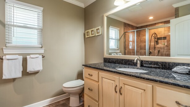 Un baño con pared color beige, inodoro blanco, lavamanos con un espejo grande arriba y zona de ducha visible a través de una puerta de cristal.